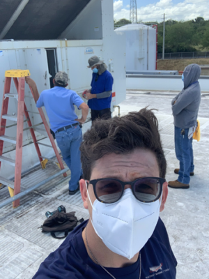 Person wearing a mask taking a selfie on a rooftop while others work on an HVAC unit in the background.